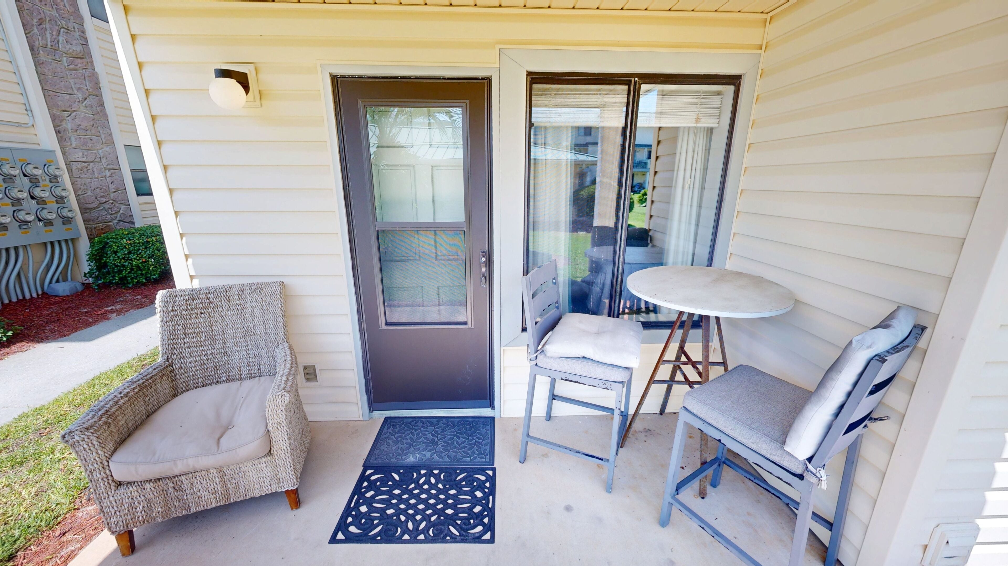 A covered porch with a door, two chairs, a small table, and a decorative mat.