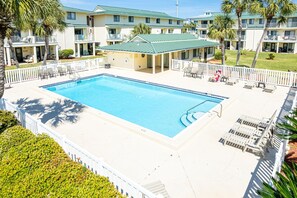 A swimming pool surrounded by lounge chairs and a small building, likely a poolside bar, with a view of hotel buildings in the background. - Sandprints B1 1 Bedroom Condo (Miramar Beach)