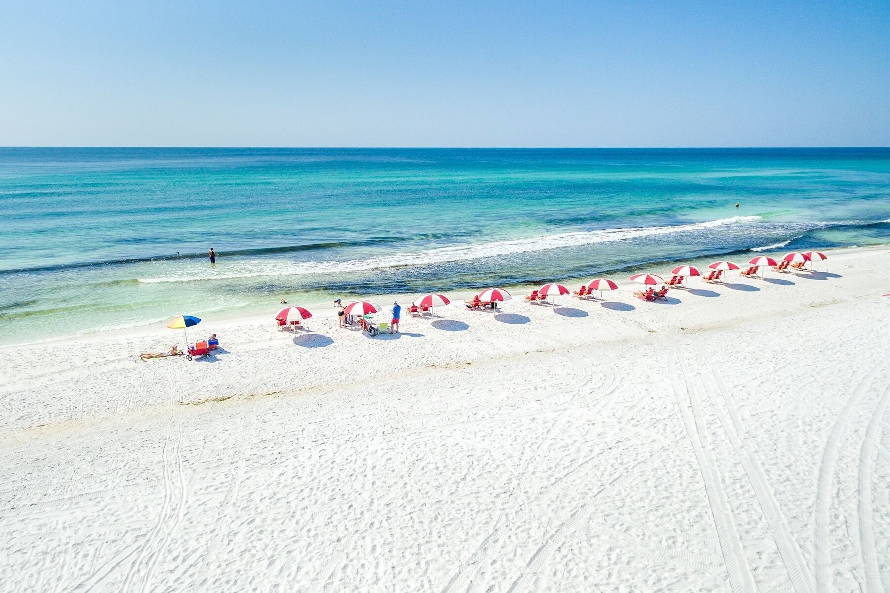 A beach with white sand, clear blue water, and red and white umbrellas.