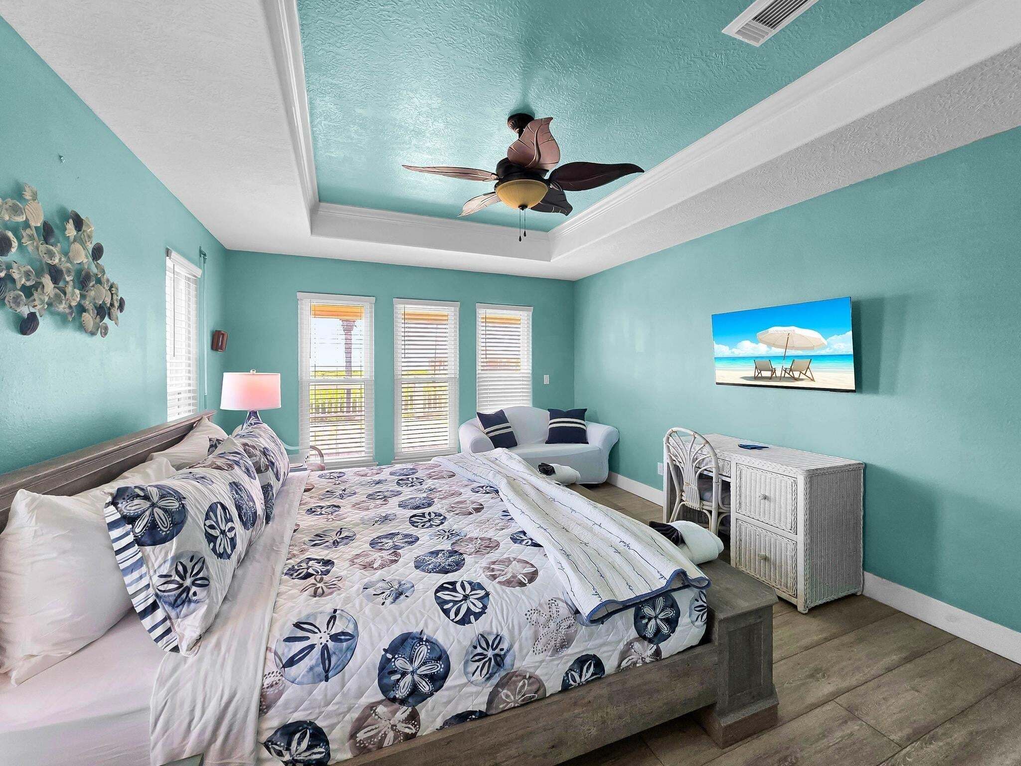 A bedroom with a bed, a chair, a table, a ceiling fan, and a beach-themed painting.