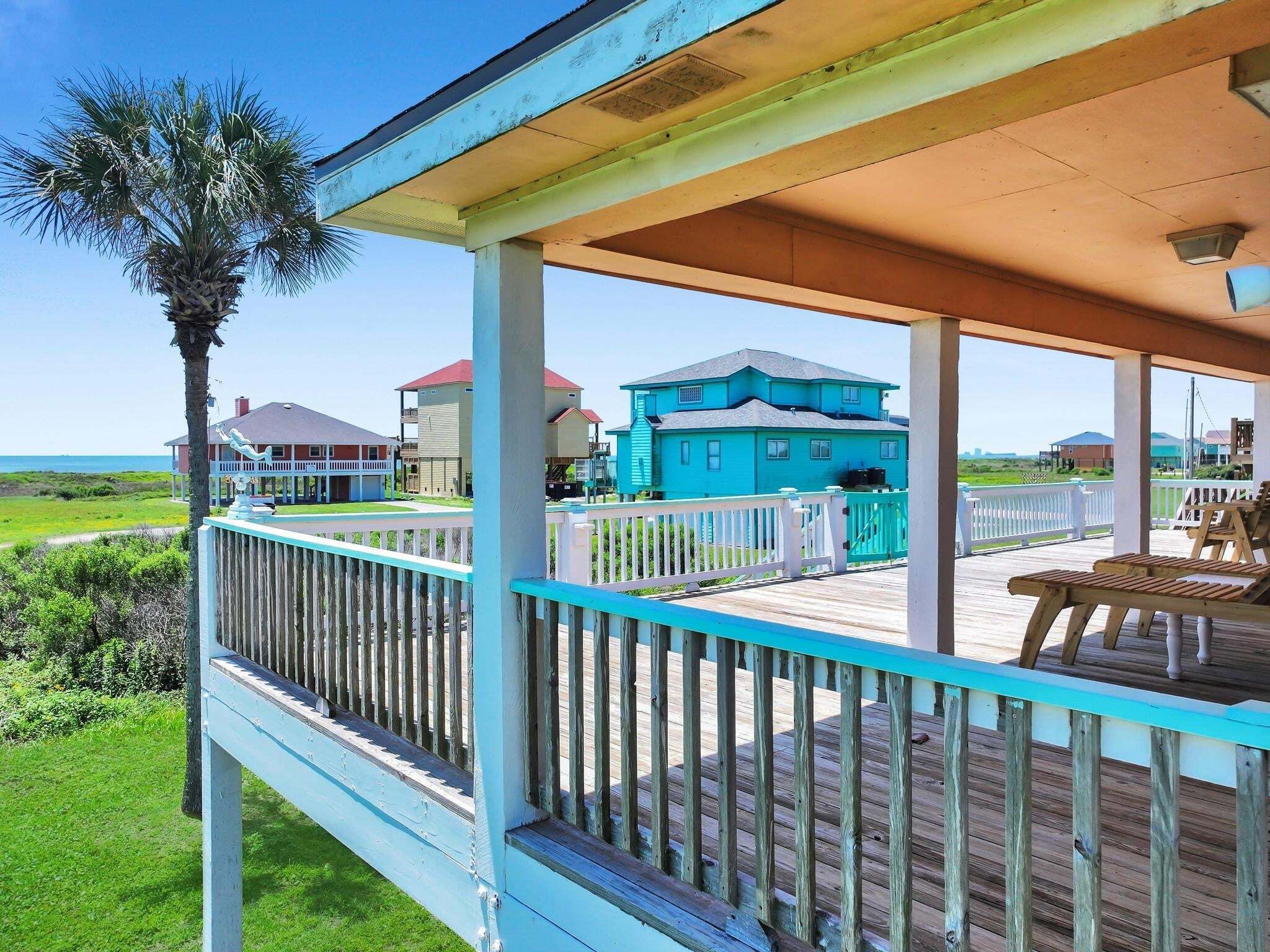 A wooden deck with a railing, a palm tree, and houses in the background.