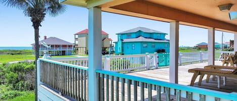 A wooden deck with a railing, a palm tree, and houses in the background.