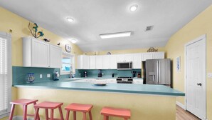 Una cocina con armarios blancos, un refrigerador de acero inoxidable, un microondas y un lavamanos. Hay tres taburetes rojos y una ventana con vistas.