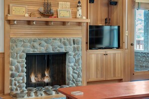 A stone fireplace with a television on a wooden stand, a shelf with decorative items, and a view of the outdoors. - Beachcomber's Retreat *Beach*Golf*Pets (Gearhart)