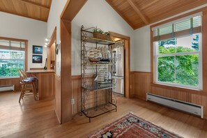 A room with wooden floors, a large window with a view of greenery, a metal shelf with decorative items, and a chair. - Beachcomber's Retreat *Beach*Golf*Pets (Gearhart)