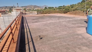 A rooftop terrace with a metal railing, a blue water tank, and a view of a town and mountains in the distance.