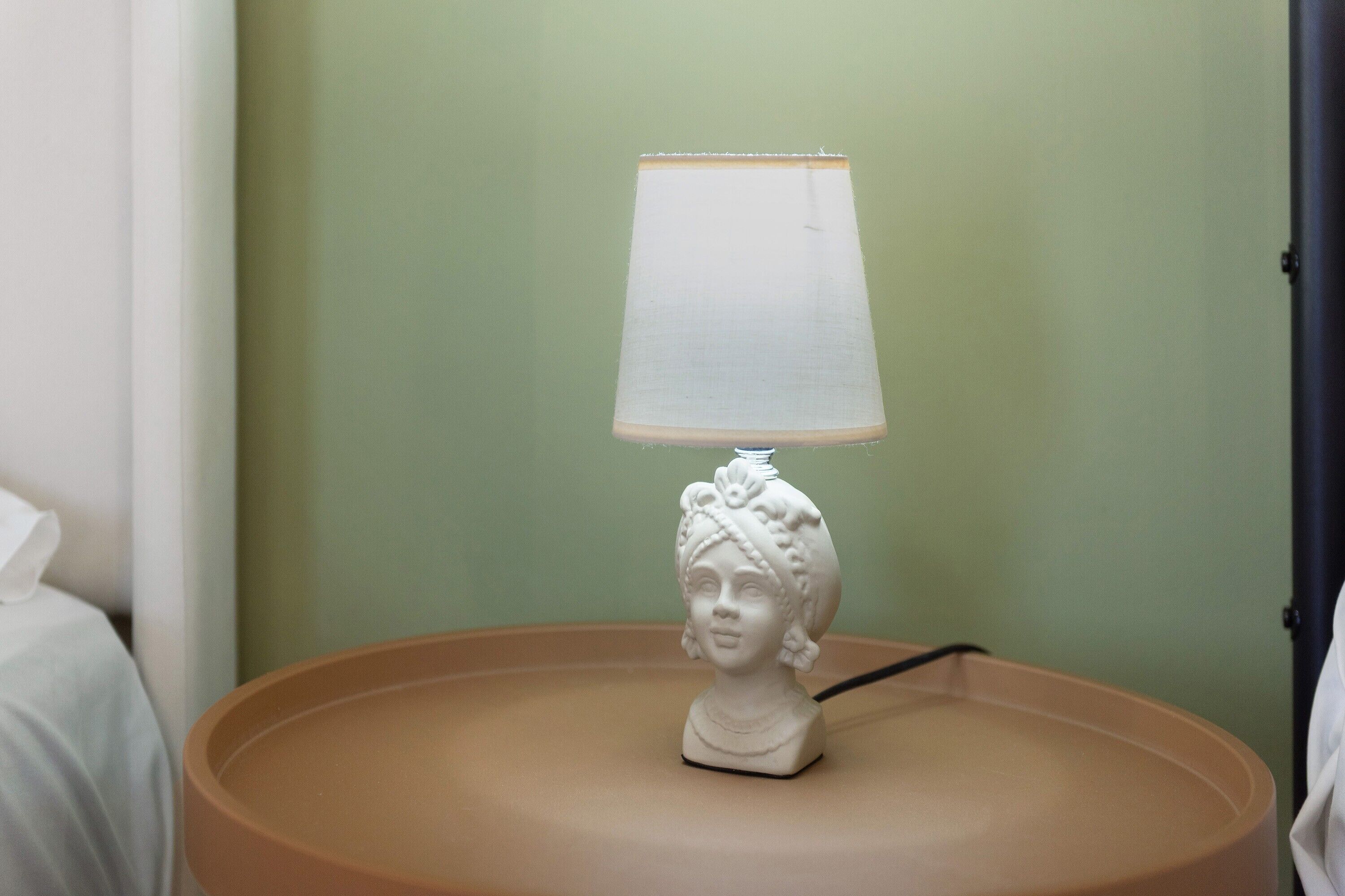 A white table lamp with a white lampshade on a round table against a green wall.