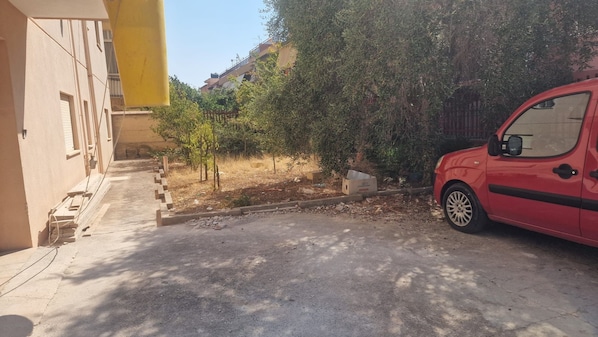 A red car parked in a driveway with a building on the left and a tree in the background.