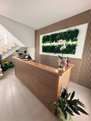 Un mostrador de recepción con detalles de madera, una pared verde con jardín vertical y una escalera con barandilla de metal.