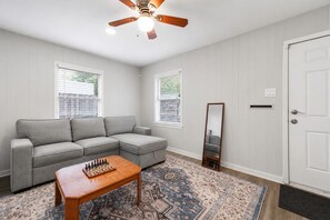 Una habitación con un sofá seccional gris, una mesa de centro de madera, un tapete estampado, un ventilador de techo y una puerta con buzón.