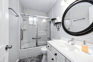 Un baño moderno con ducha compartimentada por vidrio, un lavamanos blanco con dispensador de jabón y un espejo redondo.