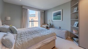 A bedroom with a large bed, a bedside table, a chair, a bookshelf, and a window with curtains. - Waterfront Luxury Apt With Pool Gym Concierge (London)