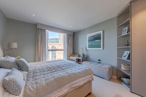 Ein Schlafzimmer mit einem großen Bett, einem Nachttisch, einem Sessel, einem Bücherregal und einem Fenster mit Vorhängen.