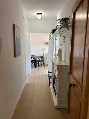 Ein Flur mit einer Holztür, einer weißen Wand mit einem gerahmten Bild, einer Deckenleuchte und einem Essbereich im Hintergrund.