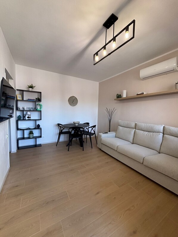Ein modernes Wohnzimmer mit Sofa, Esstisch, Stühlen und einer Küchenzeile mit Regalen und Geräten.