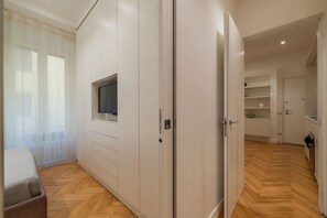 Ein modernes Zimmer mit an der Wand montiertem Fernseher, einem Kleiderschrank und einer durch eine offene Tür einsehbarer Küchenbereich.