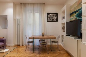 Ein modernes Wohnzimmer mit einem Holzesstisch, transparenten Stühlen, einem Glastisch, einer Couch, einem Fernseher und Kunstwerken an den Wänden.