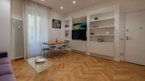 Un saló modern amb un paviment en disseny de guilló, una taula de centre de vidre, una zona de menjador amb cadires, una televisió i prestatges integrats.