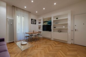 Ein modernes Wohnzimmer mit Fischgrätparkett, einem Glastisch, einer Essecke mit Stühlen, einem Fernseher und integrierten Regalen.