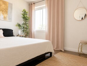 Ein Schlafzimmer mit Bett, Nachttisch, Lampe, Spiegel, Fenster und einer Pflanze.