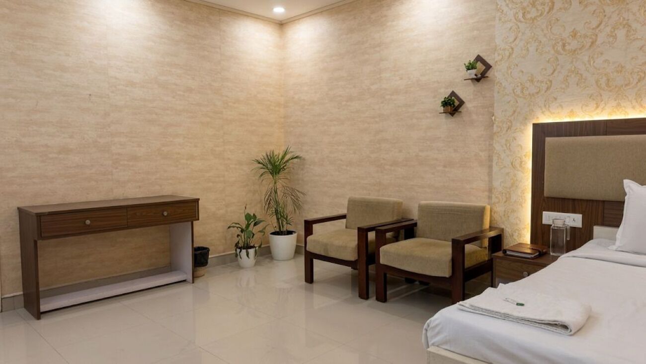 Una habitación de hotel con cama, escritorio, dos sillas, mesita de noche y dos plantas en macetas.