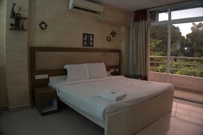 A bedroom with a large bed, a nightstand, a bedside table, a window with a view of greenery, and a wall-mounted air conditioner. - Crest Wood Inn (Kanayannur)