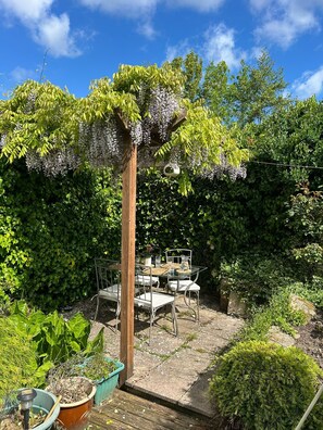 Uma pérgola de madeira com flores roxas pendentes, uma mesa com cadeiras e plantas em vasos, tudo em um jardim.