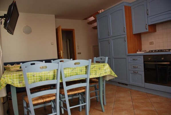 A dining area with a table set for four, blue chairs, a television mounted on the wall, and a kitchen area with cabinets and a stove. - Vrbo Property (Mastellina)