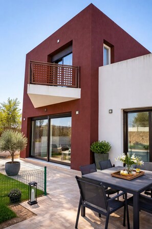 Una casa moderna de dos pisos con balcón, ventanas grandes y mobiliario para exterior.