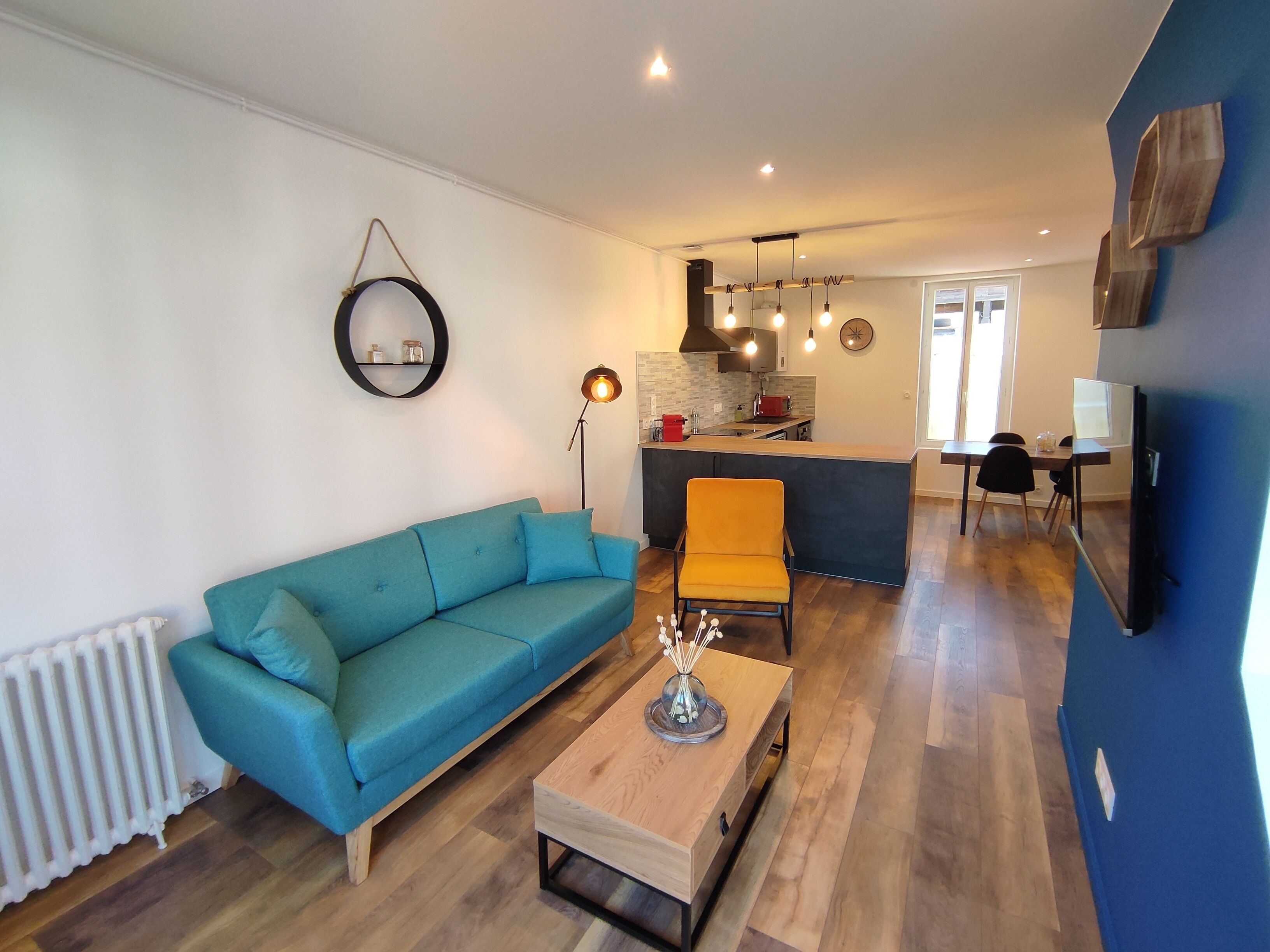 Ein Wohnzimmer mit einem blauen Sofa, einem Holztisch, einer Stehlampe und einem Küchenbereich mit Esstisch und Stühlen.