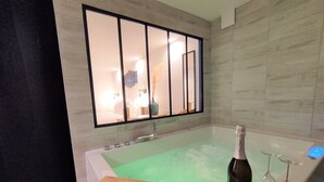 Um banheiro moderno com uma banheira grande, uma prateleira de madeira com uma garrafa e copos, e um espelho refletindo o quarto.