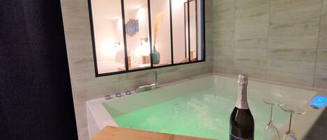 Un baño moderno con una bañera grande, un estante de madera con una botella y vasos, y un espejo que refleja la habitación.