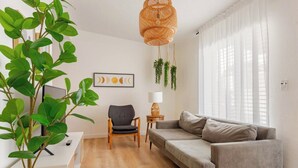 A modern living room with a grey sofa, a wooden chair, a small table with a lamp, and a large green plant. - 4BR Home Near Wynwood + Huge Tropical Yard + BBQ (Miami)