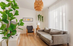 Een moderne woonkamer met een grijze bank, een houten stoel, een bijzettafeltje met een lamp en een grote groene plant.