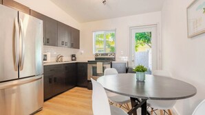 A modern kitchen with stainless steel appliances, dark cabinetry, a dining table with white chairs, and a window with a view of greenery. - 4BR Home Near Wynwood + Huge Tropical Yard + BBQ (Miami)