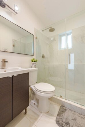 A modern bathroom with a glass shower enclosure, a white toilet, and a vanity with a mirror. - 4BR Home Near Wynwood + Huge Tropical Yard + BBQ (Miami)