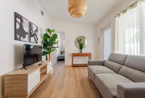 En moderne stue med et fladskærms-TV, en grå sofa, et træbord og en stor, grøn plante.