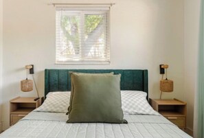 Een slaapkamer met een bed, twee nachtkastjes, een raam met zonwering en een groen gestoffeerd hoofdeinde.