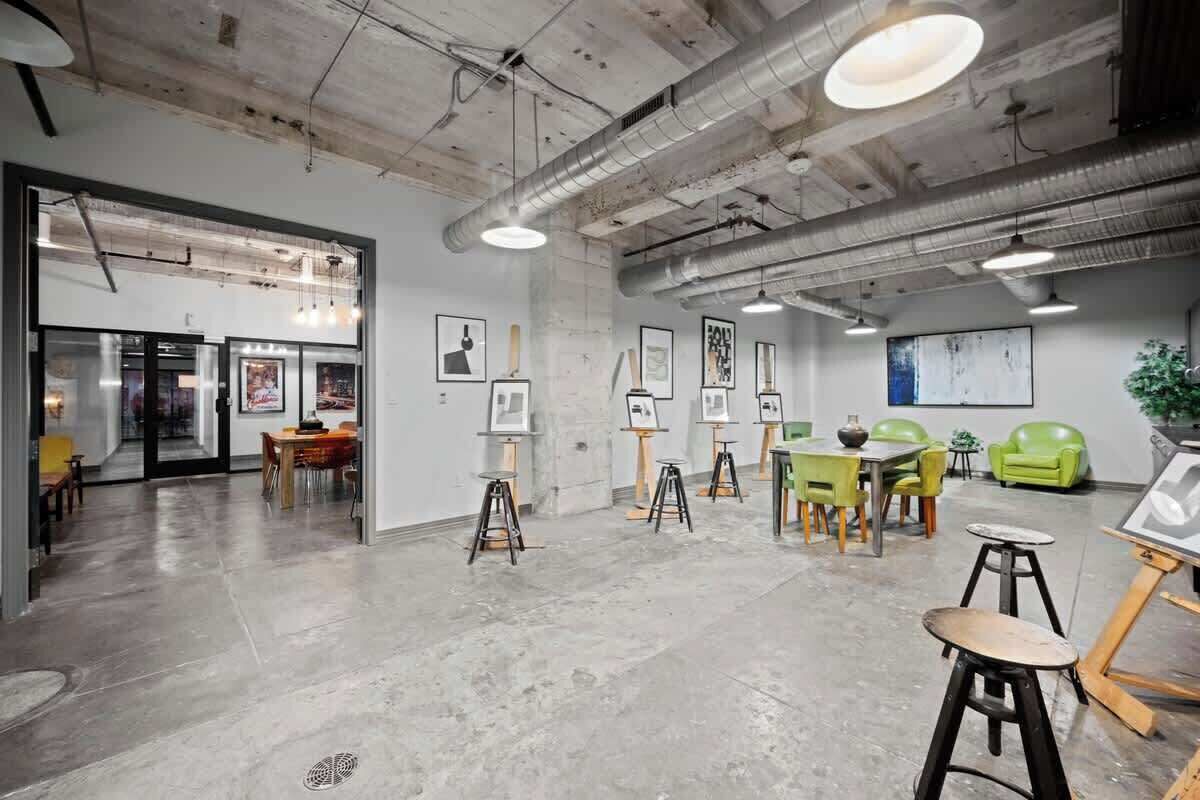 An art studio with easels, tables, and chairs, featuring a concrete floor and industrial ceiling.