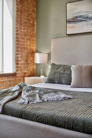 A hotel room with a brick wall, a large bed with a textured bedspread, a nightstand with a lamp, and a painting above the bed. - DT Dallas · Sleeps 12 · Pool · Gym · Views · Valet (Dallas)