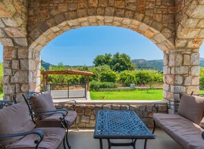 Ein Steingewölbe führt zu einer Terrasse mit einem Tisch und zwei gepolsterten Stühlen, mit Blick auf eine grüne Landschaft und Hügel.