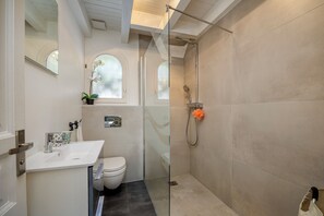 Et badeværelse med brusekabine i glas, toilet, vask og et vindue med en plante.