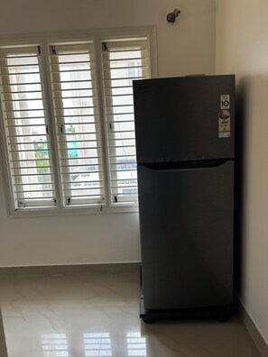 Ein Zimmer mit einem schwarzen Kühlschrank, einem Fenster mit Jalousien und einem Fliesenboden.