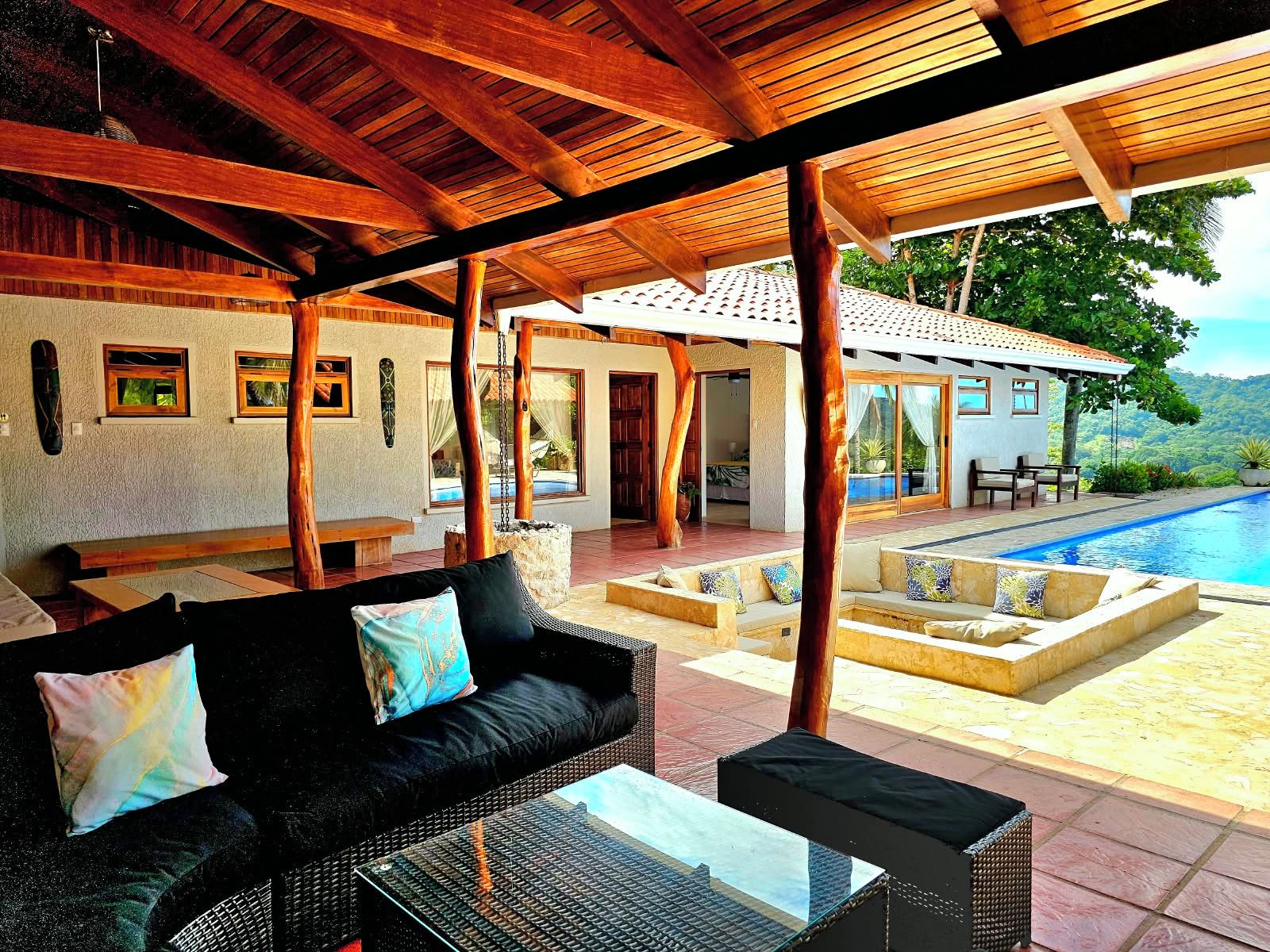 A covered outdoor seating area with a pool, wooden beams, and a view of the landscape.