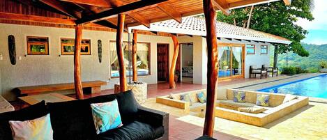 A covered outdoor seating area with a pool, wooden beams, and a view of the landscape.