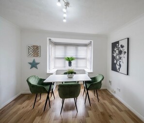 A dining area with a white table, green chairs, a potted plant, and a window with blinds. - Keepers Court Luxury Stay (South Croydon)