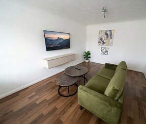 A living room with a green sofa, a round coffee table, a mounted TV, a plant, and artwork on the walls. - Keepers Court Luxury Stay (South Croydon)