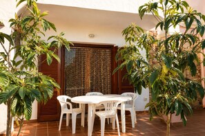 Um pátio externo com mesa redonda e quatro cadeiras de plástico branco, rodeado por plantas em vasos e uma porta de madeira com cortina estampada.