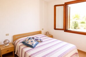 Een slaapkamer met een bed, nachtkastje, bedlampje, raam met houten kader en uitzicht op groen.