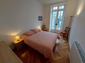 Un dormitorio con cama, lámparas de noche, una silla, un radiador y una puerta con vistas.